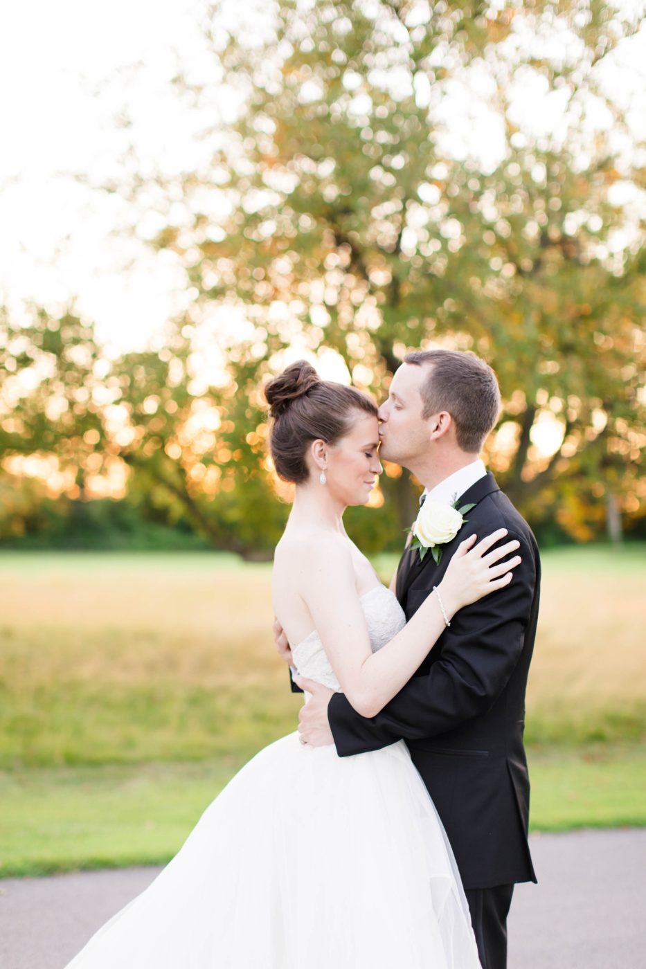 Groom giving his bride a kiss on the forehead as the sun gentle sets in the background while they stand in a field. 